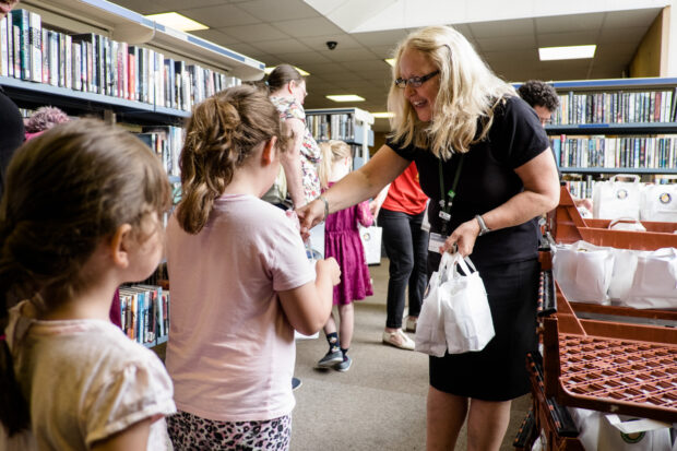 Photo of a lady handing a child a bag containing a packed lunch, in a library