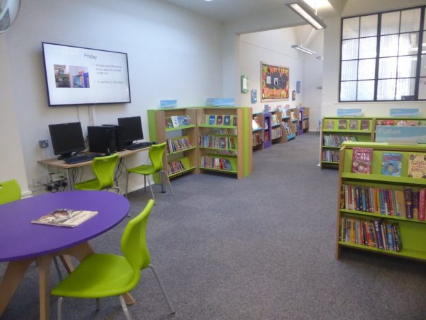 Photo of a children's section in a library which as bright green and purple furniture, lots of low bookcases and 2 computers.