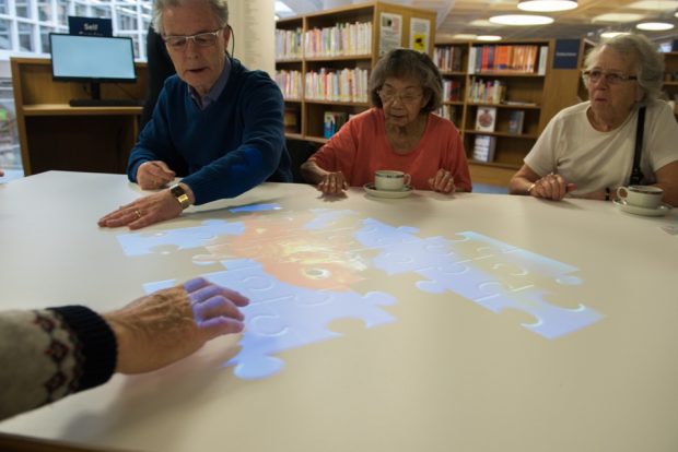 Photo of elderly people sitting around a table with a puzzle projected on it which people are doing.