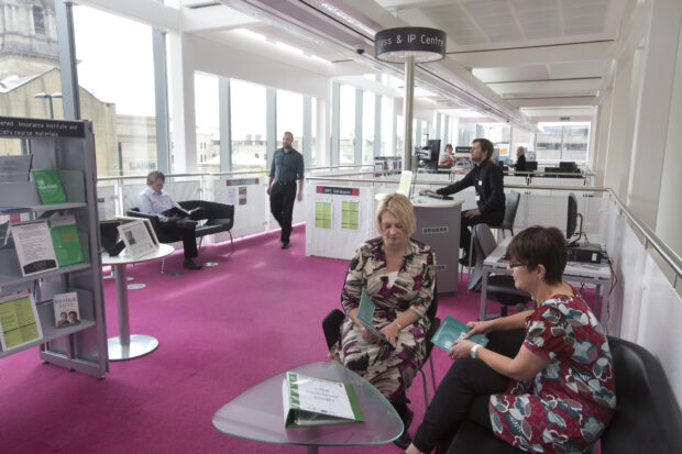 Photo of 2 women sitting at a coffee table looking at leaflets. Behind them is a man sitting at a help desk.