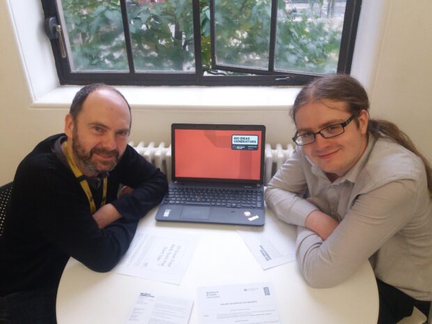 Photo of 2 men sitting at a table with a laptop between them