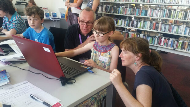 Photo of a young girl sitting at a lap top. A man and a woman watch what she's doing.