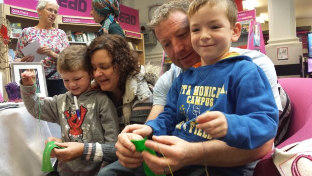 Photo of 2 young children sitting with their parents making bookmarks