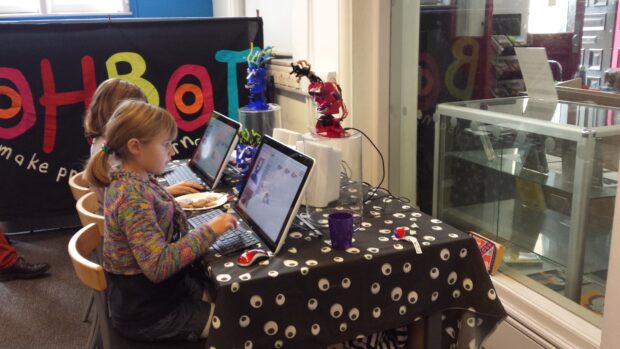 Photo of 2 children sitting at laptops and programming robotic heads which are on the table with them.