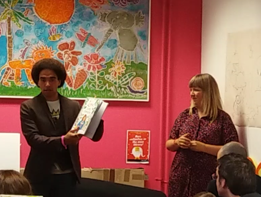 Photo of Joseph Coelho and Fiona Lumbers in Cheltenham children’s library. Joseph Coelho is standing in front of people with a book in his hand.