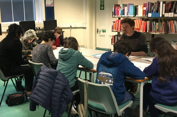 Photo of Alan working with a group of young people. Everyone is sitting down at tables and facing each other