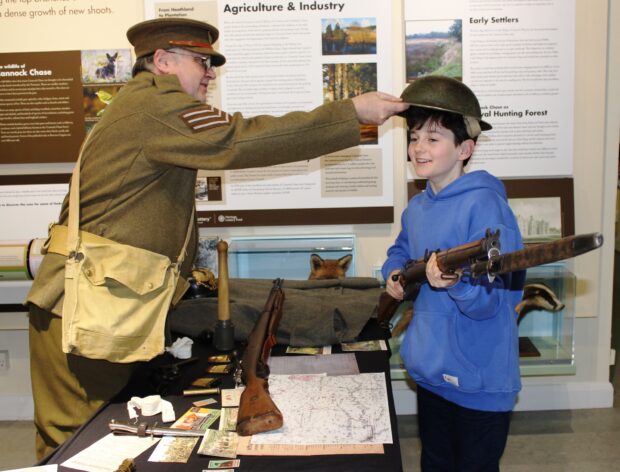 Photo of a young boy handling items in the exhibition. He is holding a rifle while an older man dressed in a soldiers uniform from World War One puts an armoured hat on his head.