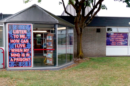 Photo of the outside of the library which has a piece of artwork in the window that says: Listen to me. How can I live when my mind is in a prison?