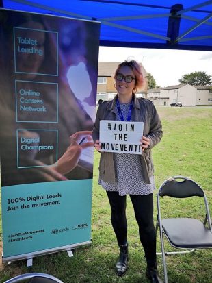 Photo of a young woman holding a sign which reads hashtag join the movement. she is standing next to a large banner at an outdoor event