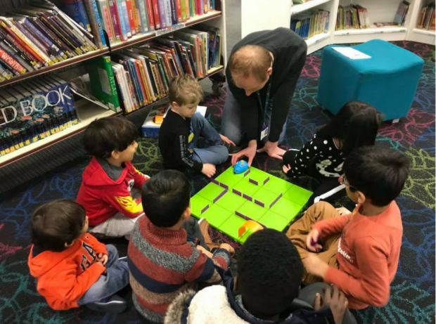 a group of young children sitting on the floor in a library, clustered around a board. an adult is sitting with them, showing them a robot about to enter a maze on the board