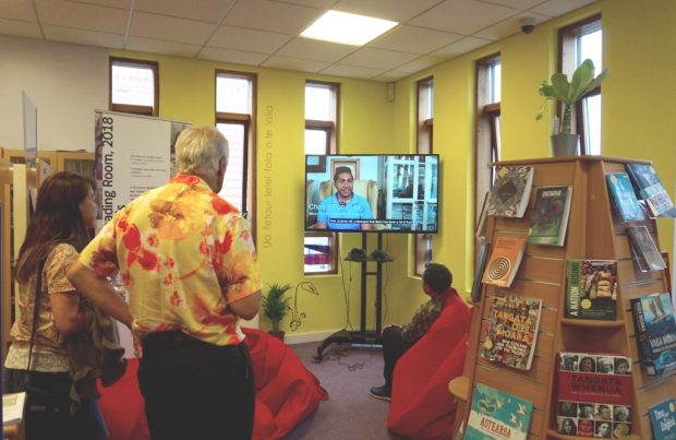 People in a corner of a library, watching a tv screen. the area is brightly coloured and some are sitting on beanbags