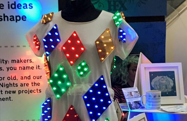 A photo of a white dress on a mannequin. On it are coloured parallelograms with LEDs on them which are lit up.