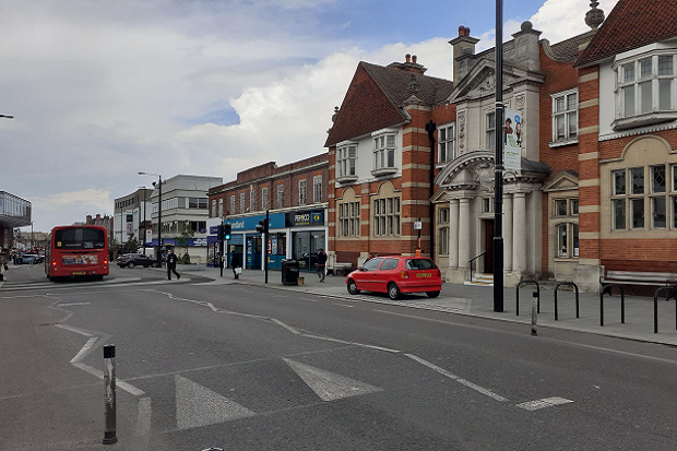 Eltham public library and shops. A red car parked outside and a bus on the road.