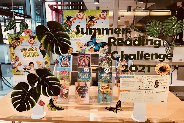 Twelve books on a clear tiered, table top, stand, behind a clear screen decorated with the words 'Summer Reading Challenge 2021', Summer Reading Challenge posters and stickers of flowers, large leaves, butterflies