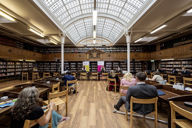 Battersea Library reading room, showing three sides of bookshelves, and gallery with second floor of bookshelves. The room has a curved glass roof, banks of wooden desks with wooden chairs. Eight audience members are sitting at the desks, looking towards the far end of the room, where Caroline Dinenage MP is speaking, flanked by two other presenters.