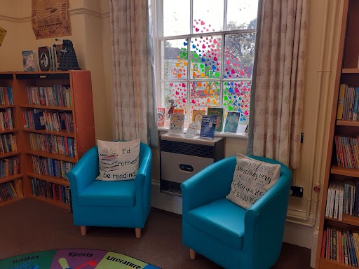 Library interior at 'The Deepings'/ Two light blue armchairs in front of window decorated with rainbow coloured heart stickers, with bookshelves on wither side